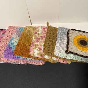 May include: A collection of seven square crochet potholders arranged in a row. The designs include a sunflower motif with a brown center and yellow petals, solid brown, variegated pink and white, solid mustard, blue and purple, and pink.