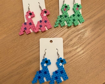 May include: Three pairs of dinosaur-shaped earrings in pink, green, and blue. Each earring is made of small, colorful beads and has a silver hook. The earrings are displayed on white cards.