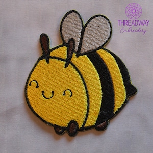 May include: Embroidered patch of a cartoon bee with a smiling face. The bee has yellow and black stripes, brown antennae and feet, and tan wings. The patch is designed for clothing or accessories. The text "THREADWAY Embroidery" is in the upper right corner.