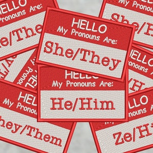 May include: A collection of red and white embroidered name tags displaying different pronoun sets such as "He/Him," "She/Her," and "They/Them." Each tag has the text "HELLO My Pronouns Are:" in a clear font, with a red border.