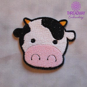 May include: Embroidered patch of a cartoon cow's face. The cow has a white face with black patches, a pink muzzle, and black eyes. The patch is outlined in black and has yellow horns. The text "THREADWAY Embroidery" is in the upper right corner.