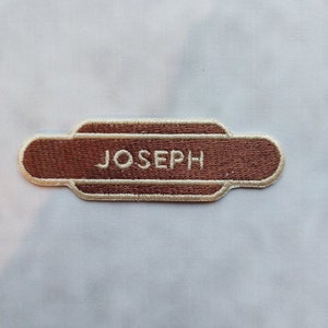May include: A brown embroidered patch with the name "JOSEPH" in white lettering. The patch has a rectangular shape with rounded ends and a decorative border. The patch is set against a white background.