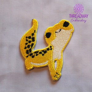 May include: Embroidered patch of a yellow gecko with black spots and white detailing. The gecko is depicted in a playful stance, with its tail curled and head tilted. The patch has a yellow border and the text "THREADWAY Embroidery".
