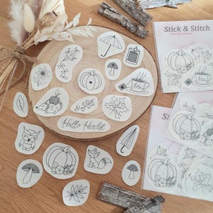 Könnte beinhalten: Ein Set mit 18 schwarzen und weißen Stickmustern mit herbstlichen Motiven, darunter Kürbisse, Pilze, Eulen, Igel, Blätter und Regenschirme. Die Muster sind auf weißem Papier gedruckt und auf einer Holzoberfläche angeordnet. Der Text "Hallo Herbst" ist in der Mitte des Bildes sichtbar.
