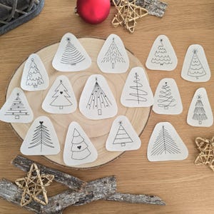 Könnte beinhalten: Eine Sammlung kleiner, weißer, dreieckiger Weihnachtsschmuckstücke mit schwarzen Strichzeichnungen verschiedener Baumdesigns. Die Ornamente sind auf einer Holzscheibe angeordnet, mit einer roten Kugel und sternförmigen Dekorationen im Hintergrund.