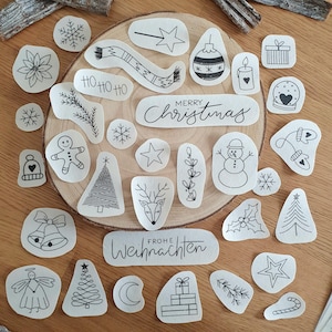 Könnte beinhalten: Eine Sammlung weißer Stoffflicken mit schwarzen Strichzeichnungen zu Weihnachten, darunter Schneeflocken, ein Schneemann, ein Weihnachtsbaum und der Text "Merry Christmas". Die Flicken sind auf einer Holzoberfläche angeordnet.