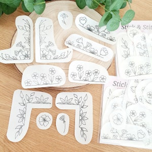Könnte beinhalten: Ein Set weißer Papierstickmuster mit floralen Designs. Die Muster umfassen Eckmuster, Bordüren und einzelne Blumen. Die Muster sind mit "Stick & Stitch" und "Stick & Stitch Embroidery" beschriftet.
