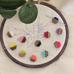 Könnte beinhalten: Eine Sammlung farbenfroher, sechseckiger Nadelmagnete aus Holz mit Akzenten in Weiß, Rosa, Gelb, Grün, Schwarz, Blau, Lila und Türkis. Die Nadelmagnete sind auf einem Stickrahmen mit floralem Design angeordnet.