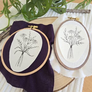 Könnte beinhalten: Zwei Stickrahmen mit floralen Designs. Einer hat einen dunkelvioletten Stoff, der andere einen hellbraunen Holzrahmen. Beide Rahmen haben einen weißen Hintergrund mit einer schwarzen Strichzeichnung eines Blumenstraußes.
