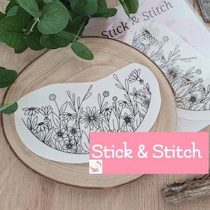 Könnte beinhalten: Ein rundes, weißes Papierstickmuster mit einer schwarzen Linienzeichnung von Blumen und Blättern. Das Muster befindet sich auf einer Holzoberfläche mit dem Text "Stick & Stitch" in rosa Buchstaben.