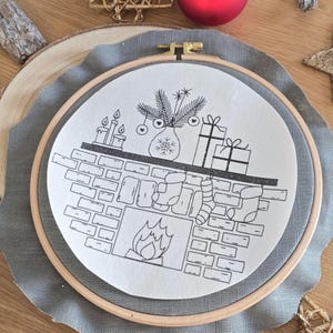 Könnte beinhalten: Eine schwarz-weiße gestickte Weihnachtsszene in einem Holzreifen. Das Design zeigt einen Kamin mit Strümpfen, Geschenken, Kerzen und einer Vase mit festlichem Grün. Die Stickerei ist auf einem weißen Stoff angebracht.
