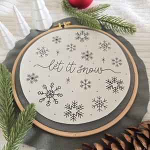Könnte beinhalten: Ein cremefarbener Stickrahmen mit dem geschwungenen Schriftzug „let it snow“ und schwarzen Schneeflocken-Designs. Der Holzrahmen liegt auf grauem Stoff, mit Kiefernzweigen und roter Dekoration im Hintergrund. Winterliches Thema.
