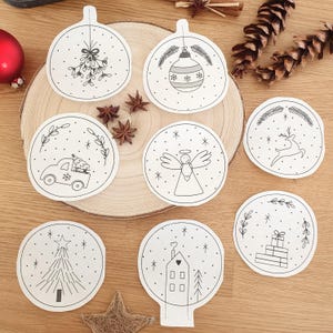 Könnte beinhalten: Acht weiße, runde Weihnachtsschmuckstücke mit schwarzen Strichzeichnungen. Zu den Designs gehören ein Auto mit einem Baum, ein Rentier, ein Engel, ein Haus, ein Weihnachtsbaum und Mistelzweige. Die Ornamente sind auf einer Holzoberfläche angeordnet.
