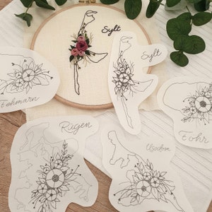 Könnte beinhalten: Fünf weiße Papierschablonen verschiedener deutscher Inseln mit floralen Designs. Die Schablonen sind mit den Namen der Inseln beschriftet: Sylt, Fehmarn, Rügen, Usedom und Föhr.