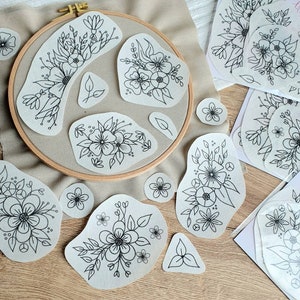 Könnte beinhalten: Ein Set mit 10 weißen Stoff-Patches mit schwarzen Linienzeichnungen von Blumendesigns. Die Patches sind auf einer hellbraunen Oberfläche angeordnet. Die Patches sind alle in verschiedenen Größen und Formen.