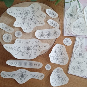 Könnte beinhalten: Ein Set mit 14 weißen Stoff-Patches mit schwarzen Blumenmustern. Die Patches sind in verschiedenen Formen und Größen, darunter Kreise, Ovale und Rechtecke. Die Designs beinhalten Blumen, Blätter und Ranken.