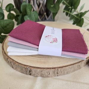 Könnte beinhalten: Ein Stapel aus vier gefalteten Stoffquadraten in den Farben Dunkelrot, Hellrosa, Grau und Weiß. Die Stoffstücke werden von einer weißen Papierbanderole zusammengehalten, die ein kreisförmiges Logo mit einem Flamingo-Motiv und Text zeigt.