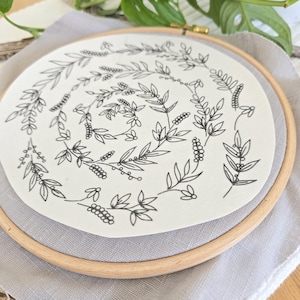 Könnte beinhalten: Ein Stickrahmen mit cremefarbenem Stoff, der eine schwarze Strichzeichnung eines botanischen Kranzes zeigt. Der Kranz enthält Blätter und Beeren. Der Holzrahmen hat einen Durchmesser von etwa 20 cm.