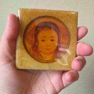 May include: A square, polished, amber-colored tile featuring a circular inset with a detailed illustration of a child's face. The tile has a smooth, glossy finish with mottled yellow and brown tones surrounding the central portrait of the child.