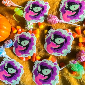 Puede incluir: Cinco pegatinas de fantasmas de dibujos animados de color morado y verde con una lengua rosa que sobresale. Las pegatinas est&aacute;n sobre una superficie amarilla peluda con ma&iacute;z dulce y otros dulces.