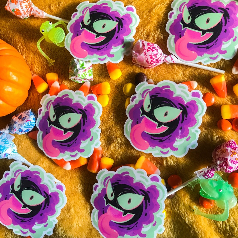 Puede incluir: Cinco pegatinas de fantasmas de dibujos animados de color morado y verde con una lengua rosa que sobresale. Las pegatinas est&aacute;n sobre una superficie amarilla peluda con ma&iacute;z dulce y otros dulces.