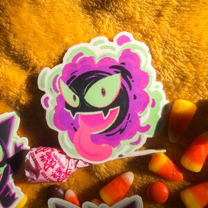 Puede incluir: Una pegatina de fantasma de dibujos animados morado y verde con una lengua rosa que sobresale. La pegatina est&aacute; sobre una superficie amarilla peluda con ma&iacute;z dulce y una piruleta.