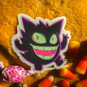 Pu&ograve; includere: Un adesivo di un fantasma di cartone animato nero e verde con un ampio sorriso pieno di denti. L'adesivo &egrave; su una superficie gialla pelosa.