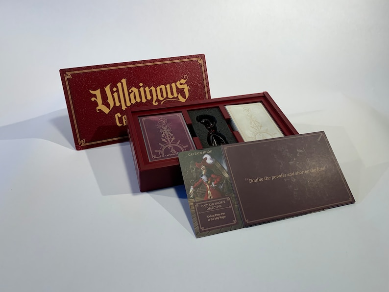 K&ouml;nnte beinhalten: Eine rote Box mit den Worten "Villainous: Cards of Darkness" auf dem Deckel. Die Box ist ge&ouml;ffnet und enth&auml;lt ein Spielbrett, Karten und eine Charakterkarte mit den Worten "Captain Hooks Ziel: Besiege Peter Pan auf der Jolly Roger" darauf.