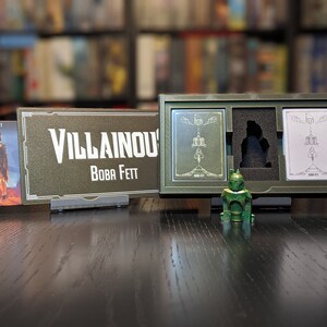 May include: A green plastic figurine of Boba Fett from the Star Wars franchise. The figurine is standing on a wooden table in front of a box with two cards inside. The box is labeled "Villainous Boba Fett".