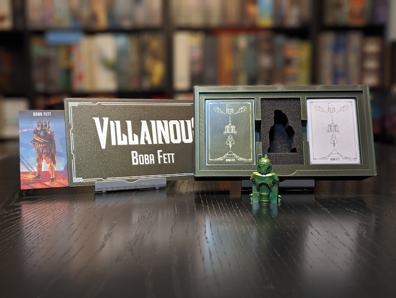May include: A green plastic figurine of Boba Fett from the Star Wars franchise. The figurine is standing on a wooden table in front of a box with two cards inside. The box is labeled "Villainous Boba Fett".