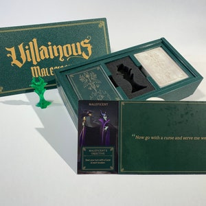 K&ouml;nnte beinhalten: Eine gr&uuml;ne Box mit den Worten "Villainous" und "Maleficent" auf dem Deckel. Die Box ist ge&ouml;ffnet und enth&auml;lt ein Spielbrett, Karten und eine Miniaturfigur von Maleficent. Die Karte lautet "Maleficents Ziel: Beginnen Sie Ihren Zug mit einem Fluch an jedem Ort."
