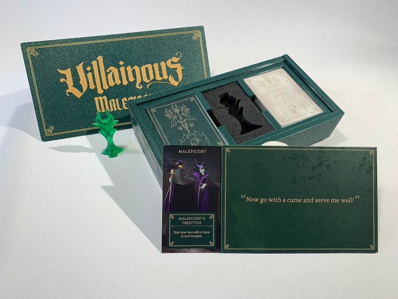 K&ouml;nnte beinhalten: Eine gr&uuml;ne Box mit den Worten "Villainous" und "Maleficent" auf dem Deckel. Die Box ist ge&ouml;ffnet und enth&auml;lt ein Spielbrett, Karten und eine Miniaturfigur von Maleficent. Die Karte lautet "Maleficents Ziel: Beginnen Sie Ihren Zug mit einem Fluch an jedem Ort."
