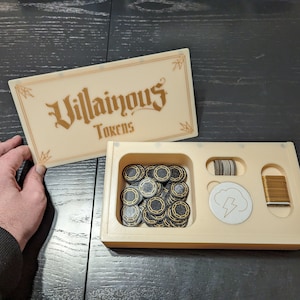 May include: A beige box with the words "Villainous Tokens" on the lid. The box is open and contains a variety of black and gold tokens, a white token with a lightning bolt, and a stack of gold-coloured metal pieces.