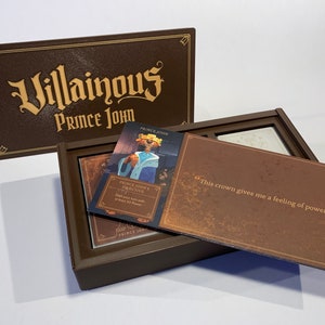 K&ouml;nnte beinhalten: Eine braune Box mit den Worten "Villainous Prince John" auf dem Deckel. Die Box ist ge&ouml;ffnet und enth&auml;lt eine Karte mit einem Cartoon-Bild von Prinz John und dem Text "Prince John's Objective: Start your turn with at least 20 Power." Die Box enth&auml;lt auch eine Karte mit dem Text "Diese Krone gibt mir ein Gef&uuml;hl von Macht!"