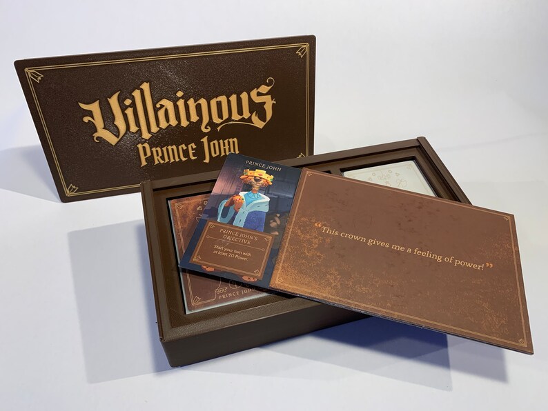 K&ouml;nnte beinhalten: Eine braune Box mit den Worten "Villainous Prince John" auf dem Deckel. Die Box ist ge&ouml;ffnet und enth&auml;lt eine Karte mit einem Cartoon-Bild von Prinz John und dem Text "Prince John's Objective: Start your turn with at least 20 Power." Die Box enth&auml;lt auch eine Karte mit dem Text "Diese Krone gibt mir ein Gef&uuml;hl von Macht!"