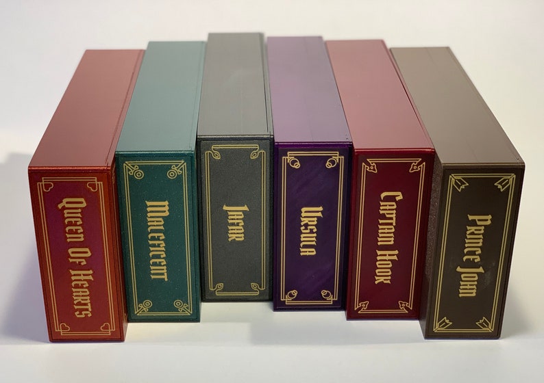 K&ouml;nnte beinhalten: Sechs dekorative Boxen mit goldenen Verzierungen und verschiedenen Charakternamen, die auf der Vorderseite aufgedruckt sind. Die Boxen sind rot, gr&uuml;n, grau, lila, burgunderrot und braun.