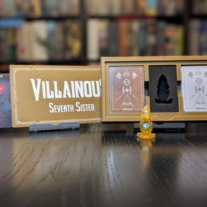 May include: A gold-colored box with a black interior and two white cards with black illustrations. The box has the text "Villainous Seventh Sister" on the front. A small gold-colored figurine is in front of the box.