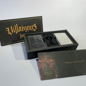 K&ouml;nnte beinhalten: Eine schwarze Box mit der Aufschrift "Villainous" in goldenen Buchstaben. Die Box ist ge&ouml;ffnet und enth&auml;lt ein Kartenspiel, eine schwarze Figur und eine wei&szlig;e Karte. Die Box enth&auml;lt auch eine Karte mit dem Text "Jafars Ziel: Stehle deinen Zug mit der Wunderlampe und sammle den Palast und die Dschinn-Marker unter deine Kontrolle."