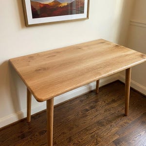 May include: A light-colored wooden desk with four rounded legs. The rectangular tabletop has rounded corners and a natural wood grain finish. The desk is approximately 48 inches long and 24 inches wide, suitable for a home office or study.