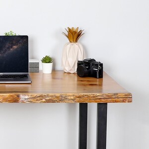 May include: A wooden desk with black metal legs, featuring a laptop, a black camera, a decorative pineapple, and a small potted plant. The desk has a natural wood grain finish.