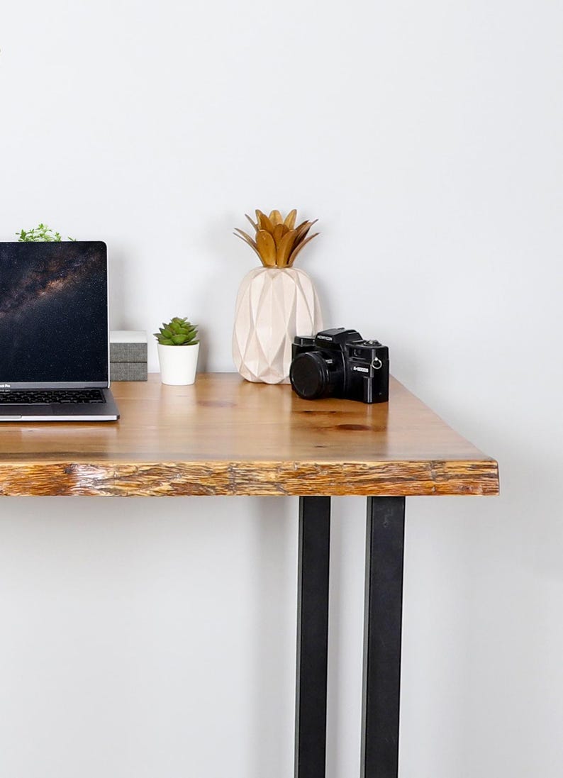 May include: A wooden desk with black metal legs, featuring a laptop, a black camera, a decorative pineapple, and a small potted plant. The desk has a natural wood grain finish.