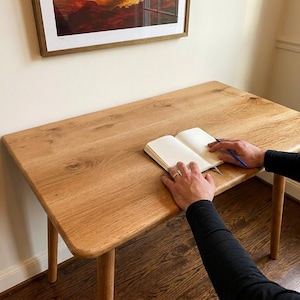 May include: A light-colored wooden desk with rounded edges and four legs. An open notebook and a pen rest on the desk's surface. The desk is positioned near a wall with a framed picture.