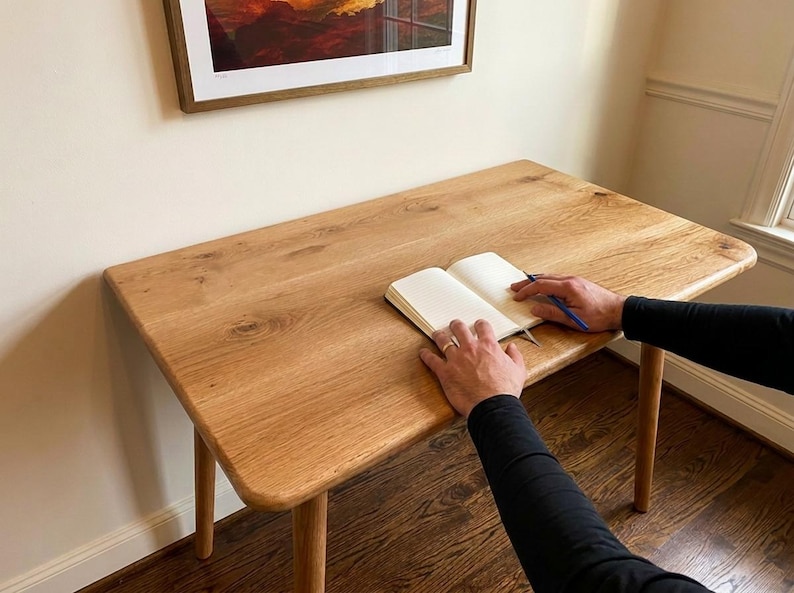May include: A light-colored wooden desk with rounded edges and four legs. An open notebook and a pen rest on the desk's surface. The desk is positioned near a wall with a framed picture.