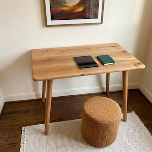 May include: A light-colored wooden desk with a tablet and a green notebook on top. A small, round, brown stool sits below the desk. The desk is supported by four angled legs. A framed painting hangs above the desk.