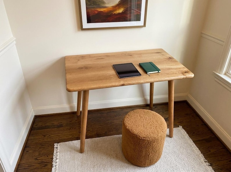 May include: A light-colored wooden desk with a tablet and a green notebook on top. A small, round, brown stool sits below the desk. The desk is supported by four angled legs. A framed painting hangs above the desk.