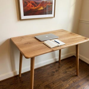 May include: A light-colored wooden desk with a rectangular top and four rounded legs. A laptop, notebook, and pen rest on the desk. A framed landscape painting hangs on the wall above the desk.