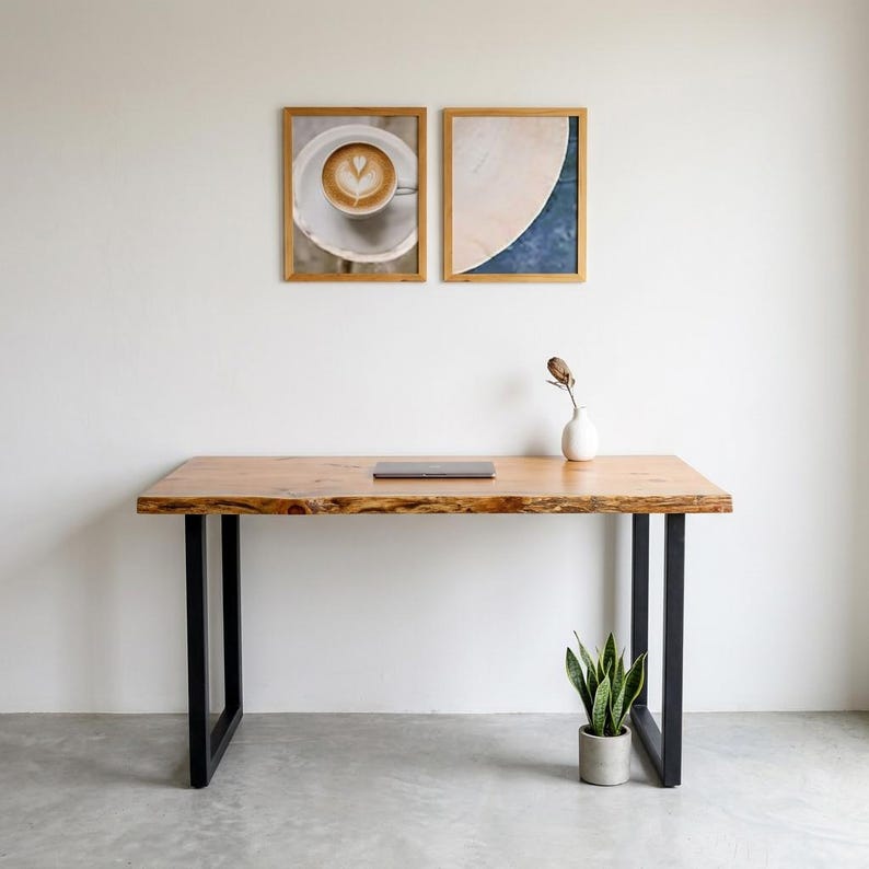 May include: A wooden desk with a live edge and black metal legs. A laptop sits on the desk. Two framed pictures hang on the white wall above the desk. A small white vase and a potted plant are on the desk.
