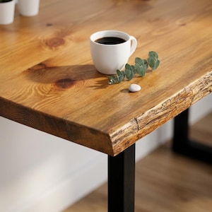 May include: A close-up of a wooden table with a natural edge, featuring a white coffee cup filled with dark liquid. A sprig of green leaves and a small white stone are placed on the table. The table has black metal legs.