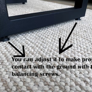 May include: Close-up of a black metal furniture leg with adjustable feet. The image shows how to adjust the feet to make proper contact with the ground using balancing screws. The leg is on a textured, neutral-colored rug.