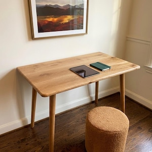 Peut inclure: Un bureau en bois clair avec des coins arrondis et quatre pieds effilés. Une tablette noire et un carnet vert sont posés sur le bureau. Un petit tabouret rond de couleur beige se trouve en dessous.